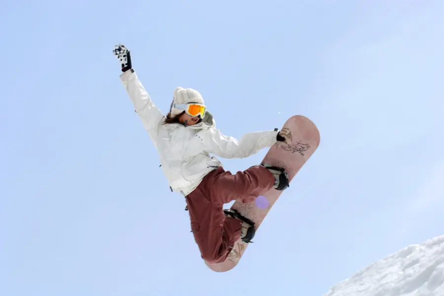 Snowboarderin springt mit Snowboard vor blauem Himmel über eine verschneite Piste.