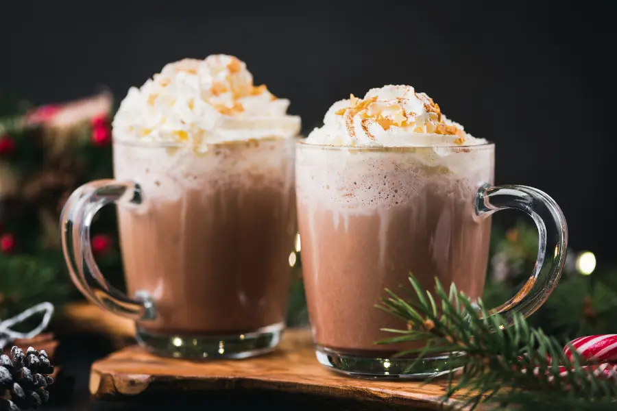 Two mugs of hot chocolate topped with whipped cream and toasted coconut, surrounded by festive greenery and holiday decorations.