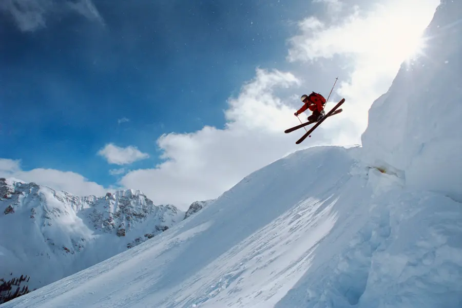 Skifahrer springt bei Sonnenschein über eine verschneite Bergkante vor blauem Himmel und alpiner Kulisse.