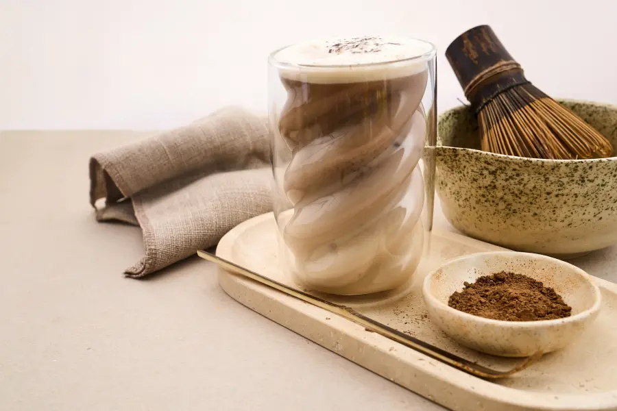 Creamy mat cha latte in a double-walled glass served with mat cha powder, a bamboo whisk, and a ceramic bowl on a neutral tabletop.