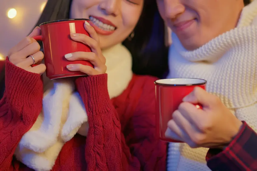 Smiling couple in cozy winter clothing holding red mugs with warm drinks in a festive, softly lit setting.