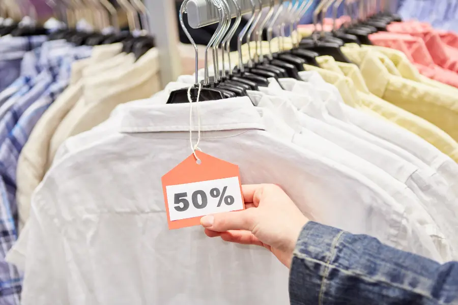 Hand hält ein 50 %-Rabattetikett an einem weißen Hemd auf einer Kleiderstange im Modegeschäft.