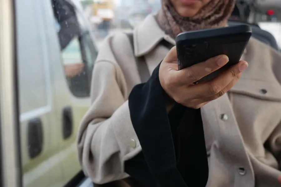 Person hält ein Smartphone in der Hand und nutzt es unterwegs neben einem Fahrzeug im städtischen Umfeld.