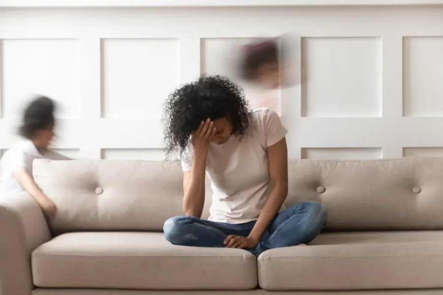 Gestresste Frau sitzt auf einem Sofa und hält sich die Stirn, während verschwommene Personen im Hintergrund Bewegung und Unruhe andeuten.