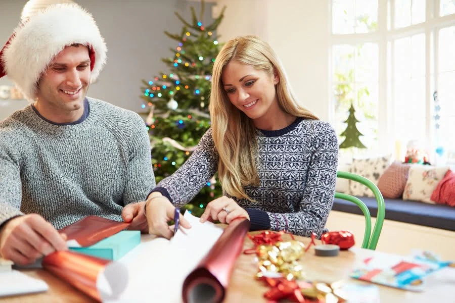 Geschenke für Kinder, Eltern, Großeltern und Freund*innen – Ideen von jung bis alt 2 Eine Frau und ein Mann sitzen an einem Tisch und packen Geschenke ein. Auf dem Tisch liegen Geschenkpapierrollen, Schleifen, Klebeband und verschiedene kleine Gegenstände. Im Hintergrund steht ein geschmückter Weihnachtsbaum, und das Zimmer wird von natürlichem Licht erhellt.