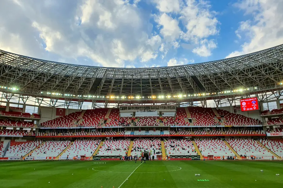 Mit dem Subside Sports Gutschein kommen Fußballt-Begeisterte voll auf ihre Kosten. Leeres Fußballstadion mit grünem Spielfeld, rot-weißen Sitzplätzen und moderner Dachkonstruktion unter blauem Himmel.