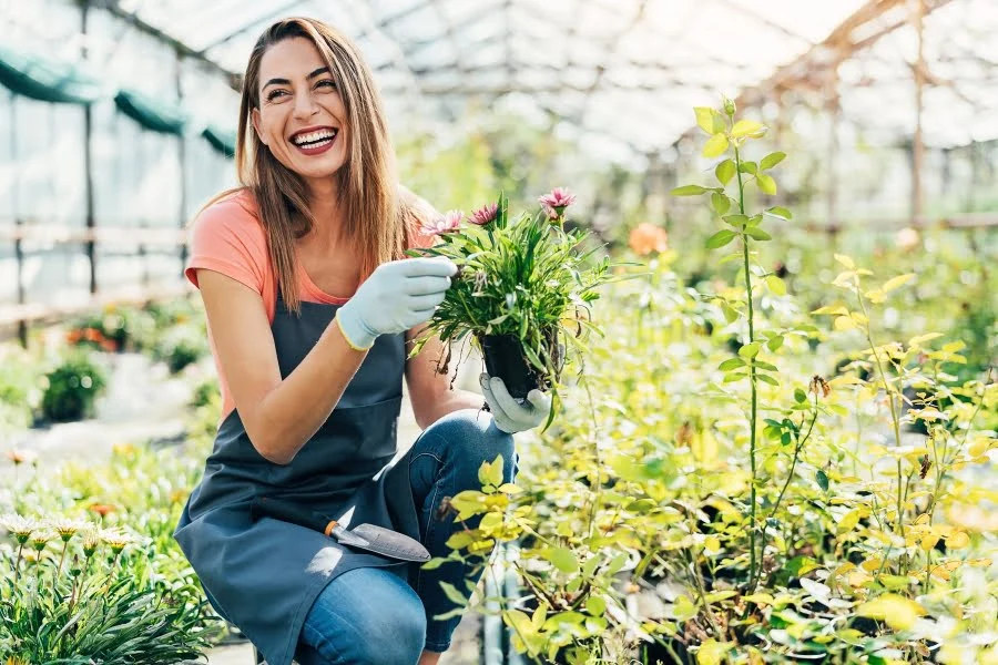 Farben, Duft und Vielfalt: So gestaltet ihr euren Garten frühlingsfit 1 Frau kniet im Gewächshaus und hält blühende Topfpflanze zwischen grünen Pflanzenreihen