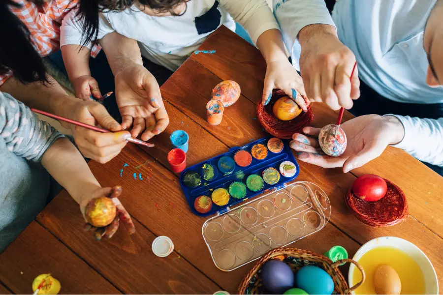 Ostern ist eine wundervolle Zeit für Kinder. Mehrere Personen bemalen gemeinsam Ostereier an einem Holztisch mit Pinseln und bunten Farben.