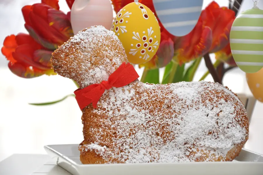 Wein ist das perfekte Getränk zu feierlichen Anlässen. Mit Puderzucker bestäubtes Osterlamm aus Kuchen auf einem Teller, dekoriert mit einer roten Schleife, vor bunten Ostereiern und roten Tulpen.