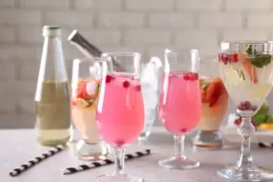 Mehrere Gläser mit bunten, alkoholfreien Cocktails in Rosa- und Gelbtönen, garniert mit Beeren, Minze und Eiswürfeln – daneben eine Flasche mit heller Flüssigkeit auf hellem Untergrund.