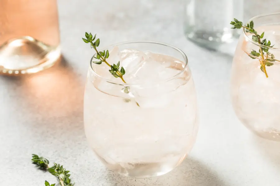 Ein Glas mit einem hellen, gekühlten Getränk auf Eis, garniert mit einem Zweig frischen Thymians – auf einem hellen Untergrund mit weiteren Gläsern im Hintergrund.