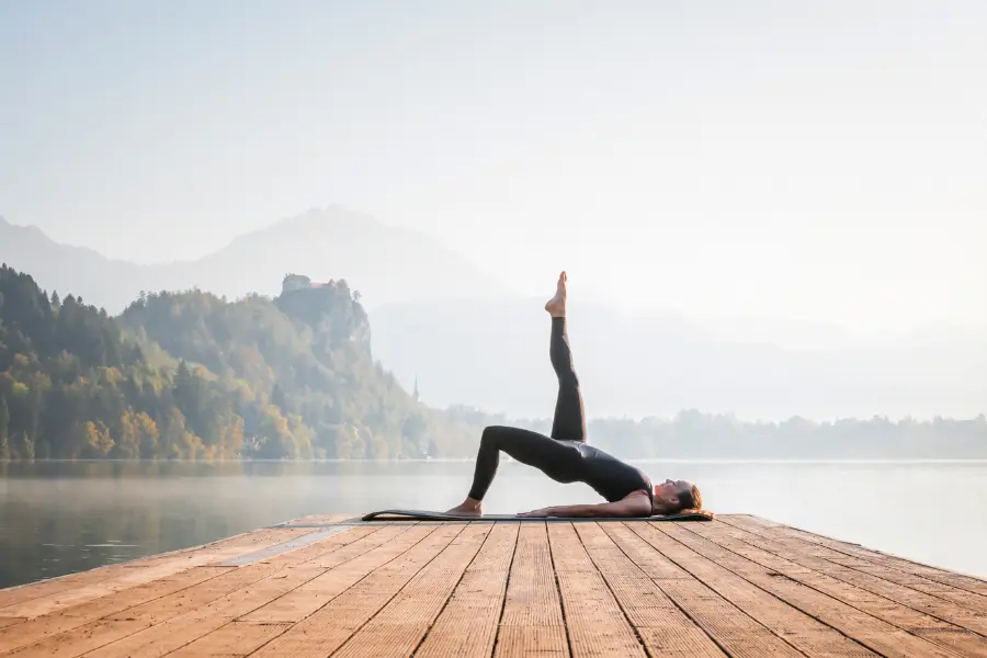 Eine Frau macht Yoga auf einem Holzsteg an einem ruhigen See, umgeben von Bergen und Morgennebel. Sie liegt auf der Matte und streckt ein Bein senkrecht nach oben – eine entspannte und kraftvolle Pose.