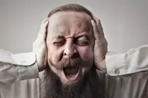 Ein bärtiger Mann mit geschlossen Augen und schmerzverzerrtem Gesicht schreit laut, während er sich mit beiden Händen die Ohren zuhält.