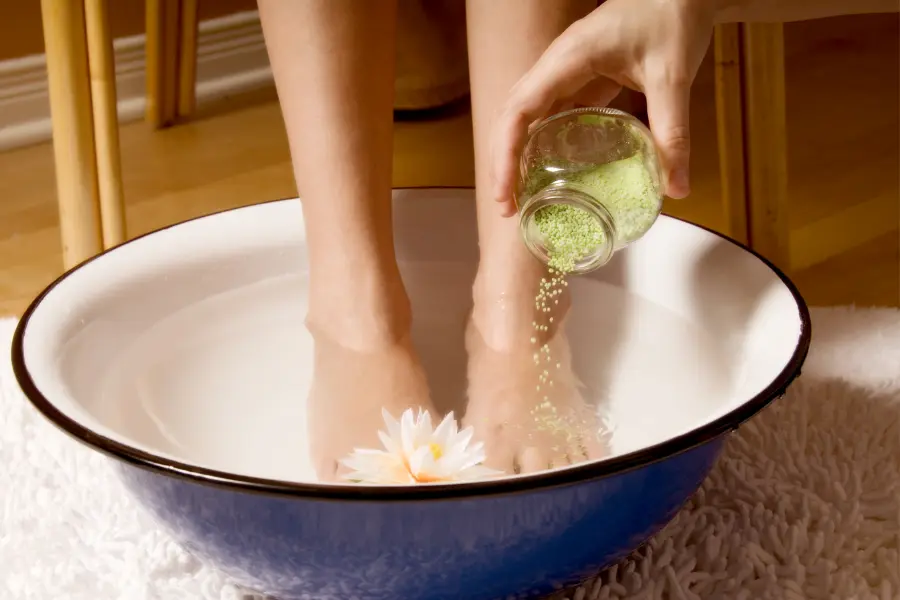 Zwei Füße in einer Schüssel mit warmem Wasser, während grüne Badesalze hineingestreut werden – daneben eine dekorative Blüte.