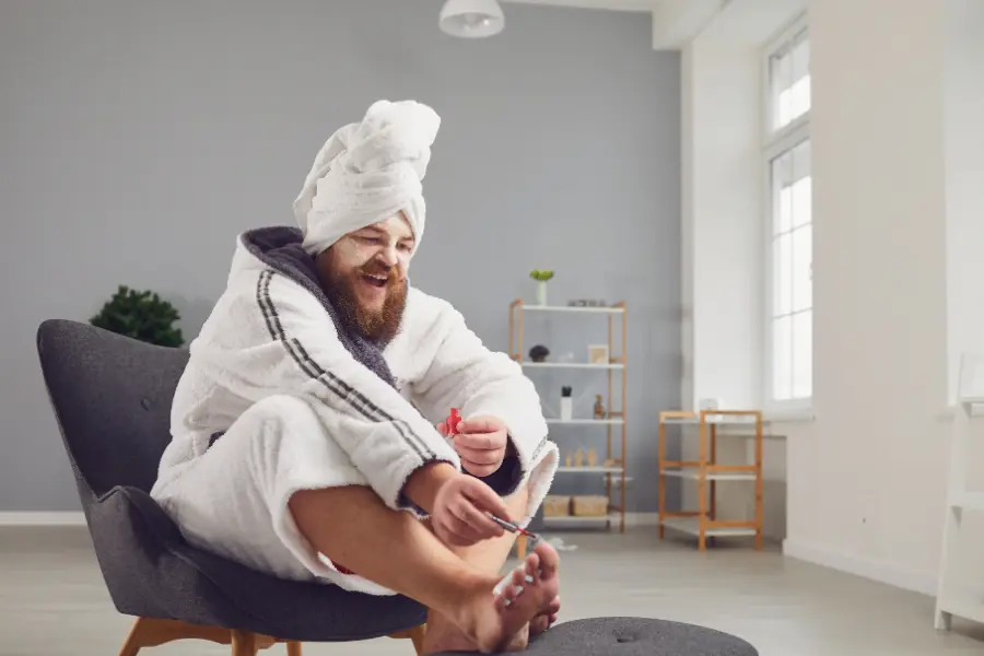 Mann in Bademantel und mit Handtuchturban sitzt entspannt auf einem Sessel und pflegt seine Fußnägel mit Nagellack.