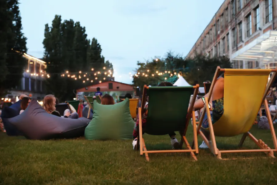 Menschen sitzen bei Einbruch der Dämmerung entspannt auf bunten Liegestühlen und Sitzkissen auf einer Wiese, umgeben von stimmungsvoller Beleuchtung, bei einem Open-Air-Kinoevent im Sommer.