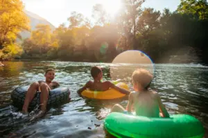 Drei Kinder treiben bei Sonnenschein lachend auf bunten Schwimmreifen in einem naturbelassenen Fluss. Im Hintergrund ist ein Wasserrad und herbstlich gefärbte Bäume zu sehen, während Sonnenstrahlen durch das Blätterdach fallen.