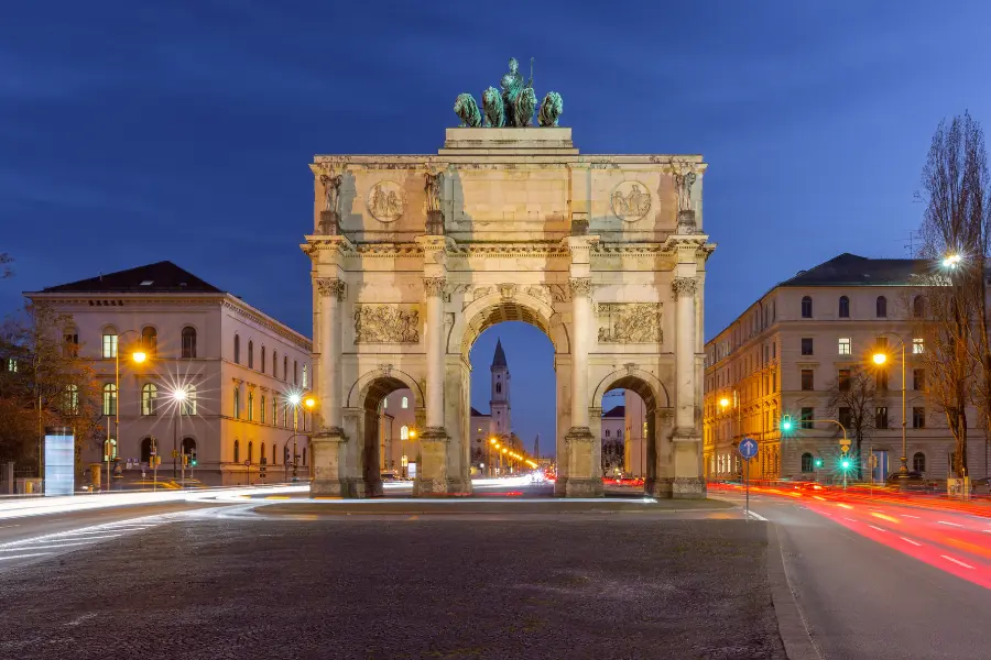Beleuchtetes Siegestor in München bei Abenddämmerung, umgeben von historischen Gebäuden und den Lichtspuren des vorbeifahrenden Verkehrs.