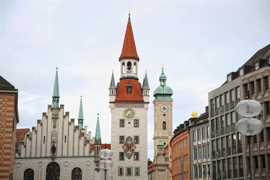 Münchens unbekannte Geschichte: Diese 3 Plätze haben Geheimnisse 1 Historischer Turm des Alten Rathauses in München mit rotem Spitzdach und kunstvoller Uhr, umgeben von Altstadtgebäuden und der Heilig-Geist-Kirche.
