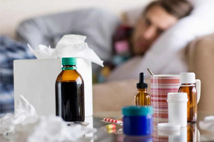 Schnelle Linderung: Wie ihr eure Erkältungsdauer verkürzen könnt – Herbst 2025 5 Im Vordergrund steht ein Glastisch mit verschiedenen Erkältungsmitteln: Fläschchen, eine Tasse mit Streifenmuster, eine Packung Taschentücher und verstreute Tabletten. Im Hintergrund liegt eine Person mit Schal auf dem Sofa und ruht sich aus. Das Bild vermittelt eine häusliche Krankensituation.