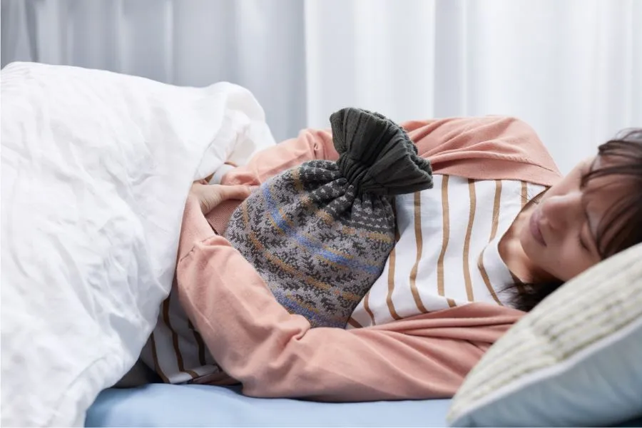 Schnelle Linderung: Wie ihr eure Erkältungsdauer verkürzen könnt – Herbst 2025 4 Eine Person liegt auf der Seite im Bett, eingehüllt in eine Decke. Sie trägt ein gestreiftes Shirt und eine rosafarbene Strickjacke. Auf dem Bauch hält sie eine Wärmflasche mit gemustertem Bezug. Der Hintergrund ist weich ausgeleuchtet, das Bett wirkt ruhig und ordentlich.