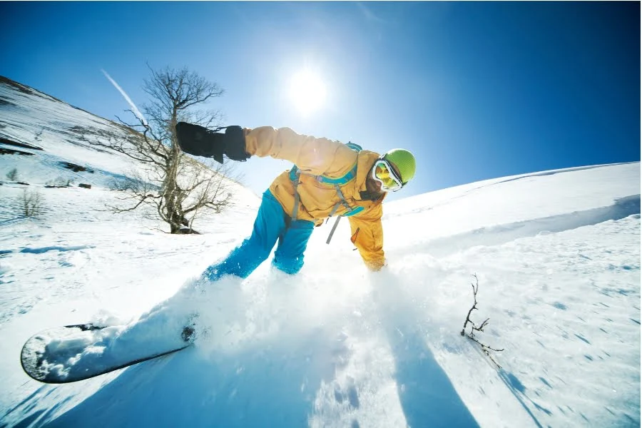 Eine Person fährt mit einem Snowboard einen verschneiten Hang hinunter. Sie trägt eine gelbe Jacke, eine grüne Helmhaube, eine Schutzbrille und blaue Schneehose. Im Hintergrund leuchtet der Himmel klar blau, die Sonne steht hoch über der Szene. Der Schnee spritzt dynamisch auf, was Bewegung und Tempo betont.