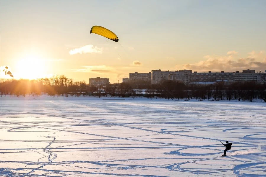 Eine Person betreibt Snowkiten auf einem zugefrorenen, schneebedeckten See. Ein großer gelber Kite zieht im Wind, während die Sonne tief am Horizont steht. Der Hintergrund zeigt eine Reihe von Gebäuden und Bäumen im warmen Abendlicht.