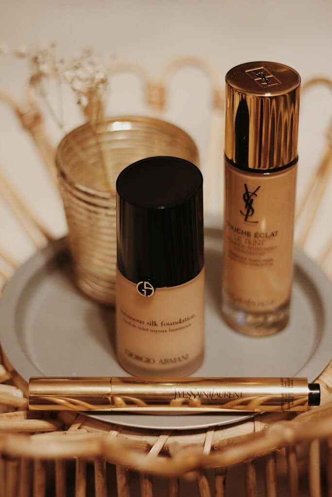 Auf einem Teller stehen Foundations von Giorgio Armani und Yves Saint Lauren, sowie ein Concealer. Geschmückt wird das Bild mit Blumen im Hintergrund. 