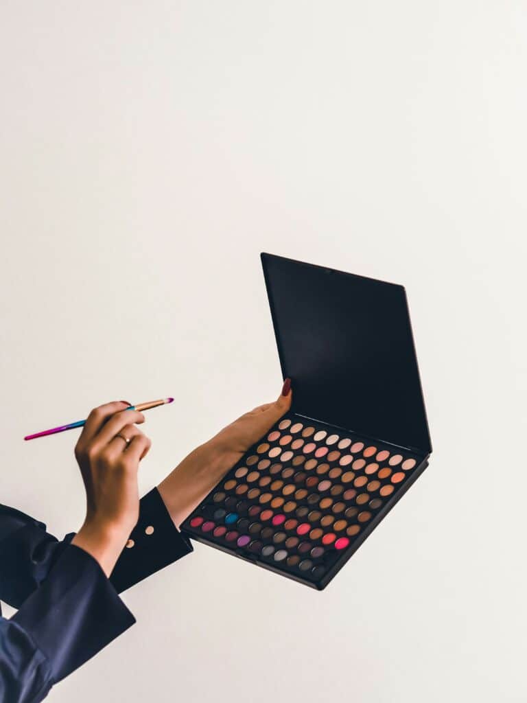Lidschattenpalette für Augenmakeup mit bunten Farben für ein Sommerliches Augenmakeup. Große Palette wird von einer Damenhand in der Hand gehalten, mit Pinsel in der anderen Hand.