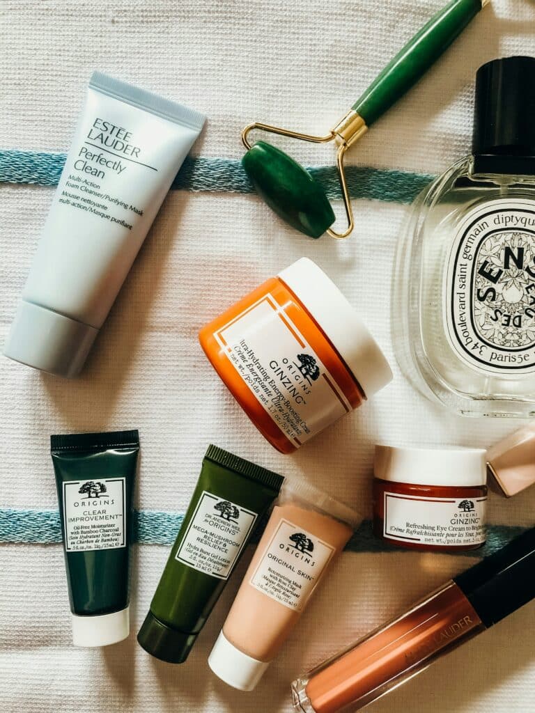 Hautpflegeprodukte, wie von Estee Lauder und origins, sowie einen Gesichtsroller, Parfum und ein Lipgloss, liegen verteilt auf einer Decke. 
