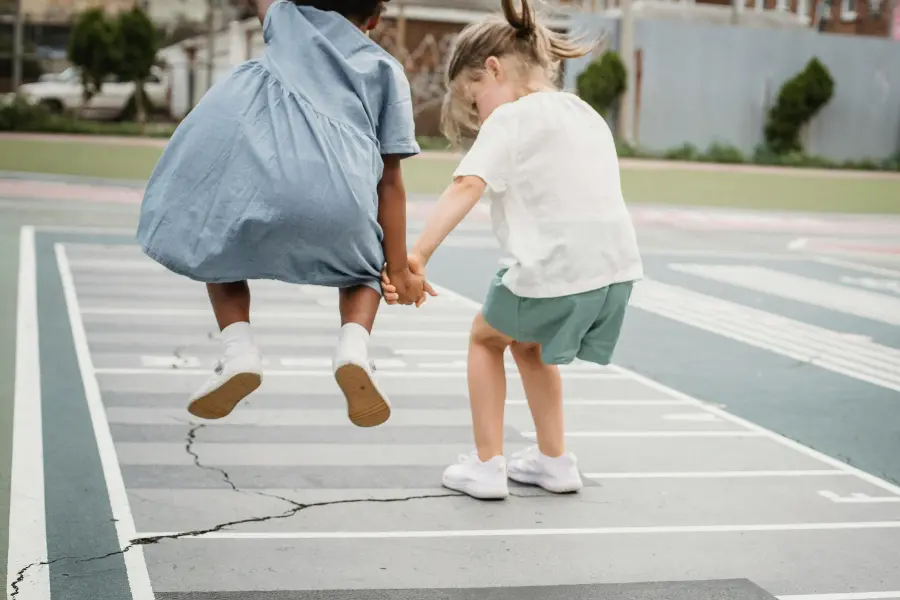 Zwei Kinder springen auf einem Spielfeld im Freien, Hand in Hand, lachend und voller Bewegung – Momentaufnahme kindlicher Lebensfreude und echter Begegnung.