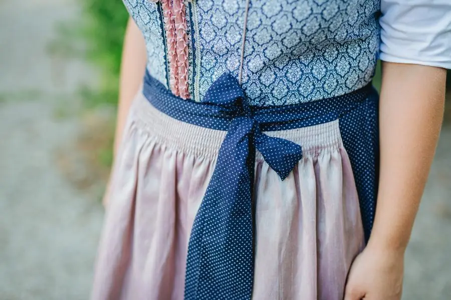 O'zapft is! - das richtige Outfit zum Oktoberfest 2025 2 Zu sehen ist der Mittelteil eines klassischen Dirndls mit rosafarbener Schürze und gemustertem Mieder in Blau. Die Schleife ist links gebunden – traditionell ein Hinweis darauf, dass die Trägerin ledig ist. Das Bild zeigt keine Person im Ganzen, sondern fokussiert auf das Detail der gebundenen Schürze.