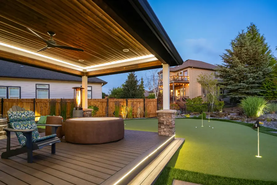 Hausbauen ist mit dem Musterhaus Gutschein ganz einfach. Ein stilvoll gestalteter Gartenbereich mit überdachter Terrasse, LED-Beleuchtung, Whirlpool und Putting-Green, eingerahmt von modernen Häusern und gepflegten Pflanzen.