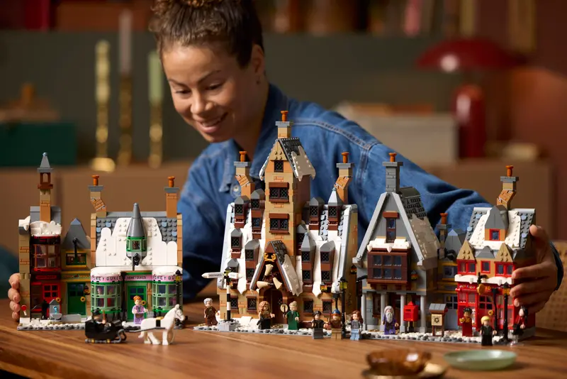 LEGO begeistert im Herbst – Von Hogwarts bis zur IAA – Die spannendsten Neuheiten im Überblick 2 Frau spielt mit dem LEGO Harry Potter Hogsmeade Set auf einem Holztisch. Die verschneiten Häuser und Minifiguren wie Harry, Hermine und Ron sind detailreich aufgebaut und versetzen in die magische Welt von Hogwarts. Die Szene ist warm beleuchtet und zeigt Begeisterung für kreativen Bauspaß.