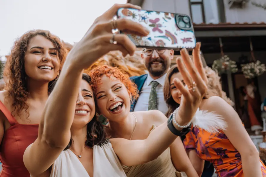 Gruppe junger Menschen, die draußen bei Tageslicht fröhlich ein Selfie mit einem Smartphone machen.