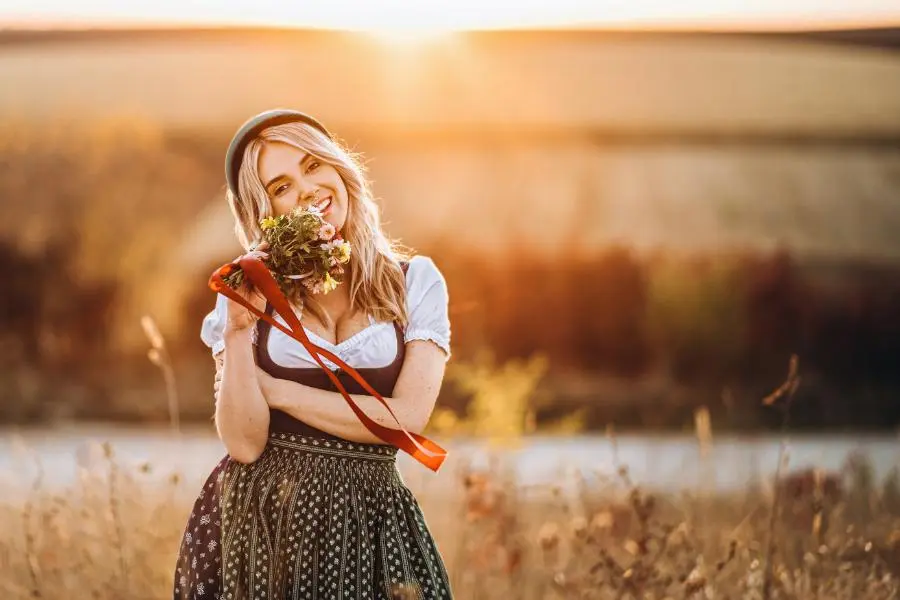 O'zapft is! - das richtige Outfit zum Oktoberfest 2025 4 Frau im Dirndl mit Blumenstrauß auf einem Feld bei Sonnenuntergang – bayerische Tradition und Lebensfreude im Spätsommer.