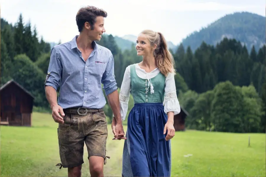 Paar in Tracht spaziert durch Berglandschaft – Lederhose und Dirndl mit mittig gebundener Schleife in traditioneller Umgebung.
