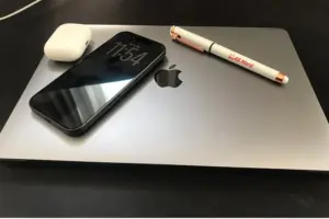 Auf einem geschlossenen MacBook liegt ein schwarzes iPhone mit aktiviertem Always-On-Display, das die Uhrzeit anzeigt. Daneben befinden sich ein weißes Ladecase für AirPods und ein Kugelschreiber mit 45 Nord-Logo. Die Geräte sind auf einer dunklen Oberfläche arrangiert, das Apple-Logo des MacBooks ist gut sichtbar.