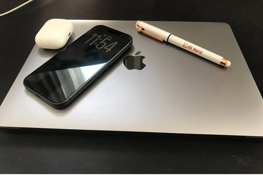 Auf einem geschlossenen MacBook liegt ein schwarzes iPhone mit aktiviertem Always-On-Display, das die Uhrzeit anzeigt. Daneben befinden sich ein weißes Ladecase für AirPods und ein Kugelschreiber mit 45 Nord-Logo. Die Geräte sind auf einer dunklen Oberfläche arrangiert, das Apple-Logo des MacBooks ist gut sichtbar.