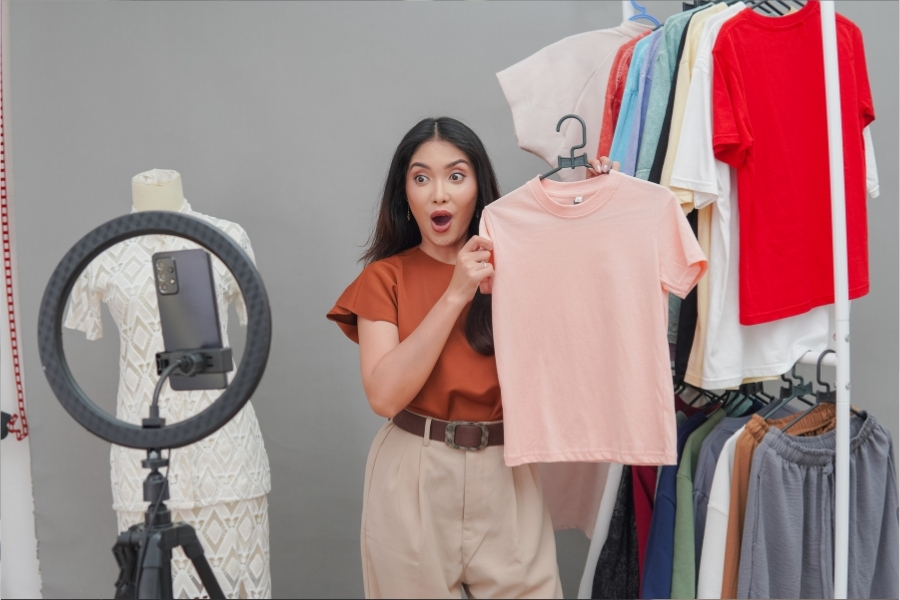 Live Shopping im Aufschwung: Wie Livestreams den Onlinehandel verändern 2 Frau präsentiert T Shirt vor Smartphone Kamera und Ringlicht in einem Raum mit Kleiderständer