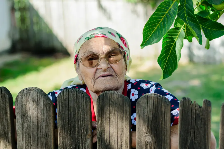 Balkon oder Garten – So gestaltet ihr euer privates Paradies 1 Ältere Frau mit Kopftuch und Brille lehnt an einem Holzzaun im Garten und blickt freundlich in die Kamera.