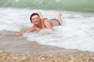 Lächelnde Frau liegt in einem Bikini am Strand im flachen Meerwasser, während sanfte Wellen sie umspülen.