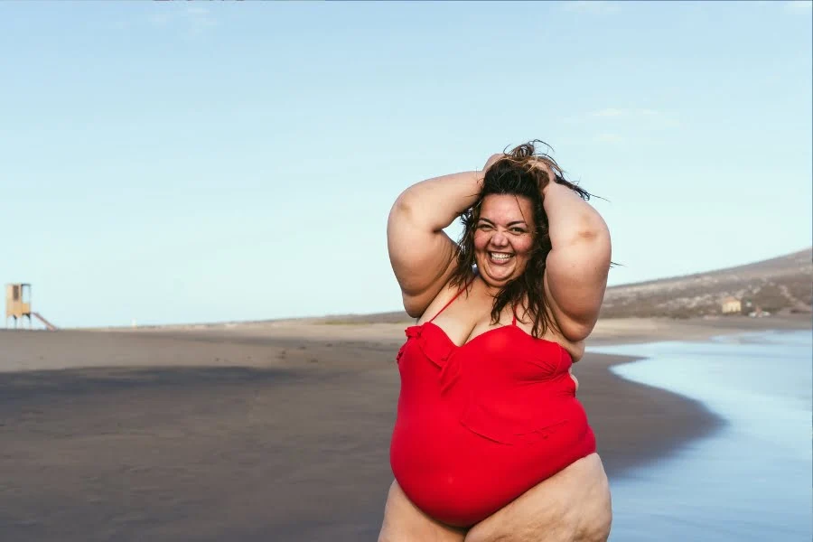 Bikini-Modetrends - das sind die absoluten Hingucker Modelle! 3 Lächelnde Frau in rotem Badeanzug steht selbstbewusst am Strand vor Meer und hellem Himmel.