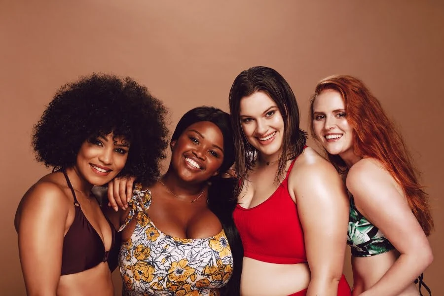 Bikini-Modetrends - das sind die absoluten Hingucker Modelle! 1 Vier Frauen in farbenfroher Bademode posieren lächelnd vor einem warmen Studiohintergrund.